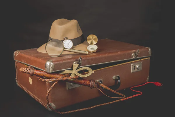 Reise- und Vorteilskonzept. Vintage-brauner Koffer mit Uhr, Fedora-Hut, Ochsenziemer, Kompass, Lupe und Ankh-Schlüssel des Lebens auf dunklem Hintergrund Stockfoto