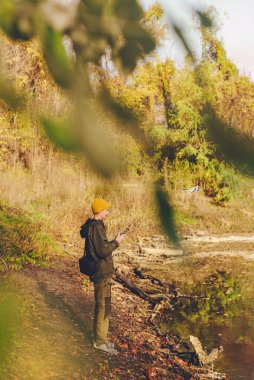 Gölün kıyısında ya da gölette duran genç bir adam hava keşfi için dron kumandası tutuyor. Sonbahar manzarasının fotoğraflarını ve videolarını çekmek için deniz kenarında insansız hava aracı kullanan biri. Arka planda uçan İHA.