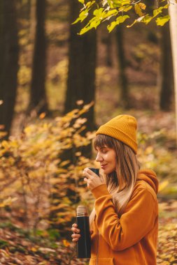 Sonbahar ormanında sıcak içecekten hoşlanan bir kadın. Yüzüne bardak tutan kadın termostan sakin bir ifadeyle su kokluyor ya da yudumluyor. Kız dinlenmek için zaman ayırıyor, dışarıda vakit geçiriyor..
