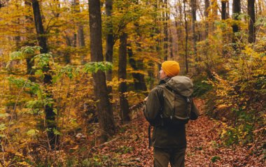 Sarı bereli, turist sırt çantalı bir gezgin, sonbahar patikasındaki yaprağın üzerindeki kameradan uzaklaşıyor. Aktif erkek yürüyüşü, açık hava macerası ya da sonbahar aylarında eğlence yürüyüşü.