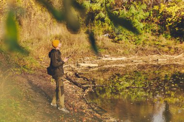 Göl kıyısında drone kontrolörü tutan bir gezgin. Sonbahar manzarasının fotoğraflarını ve videolarını çekmek için deniz kenarında insansız hava aracı kullanan biri. Suyun üzerinde sonbahar yapraklarıyla uçan drone..