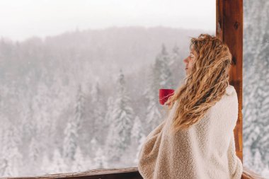 Kır evindeki karlı orman zemininde, ekose battaniyeye sarılı, bir fincan sıcak içecekle sakin bir kış sahnesi. Kadın huzurlu bir kış kaçamağı ve rahat bir rahatlama yaşıyor..