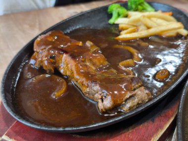 Sıcak dökme bir tabakta nefis cızırtılı biftek, üzerine bol kahverengi sos ve soğan, yanında altın patates kızartması ve taze yeşil brokoli servis edilir.