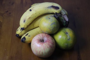 Kahverengi benekli olgun muzlar, taze bir elma ve iki portakal, klasik bir natürmort kompozisyonu içinde ahşap bir masada yan yana otururlar.