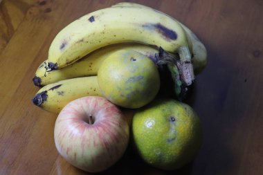 Tahta bir yüzey üzerinde olgun muzlar, bir elma ve iki mandalina portakalı bulunan taze meyve çeşitleri.