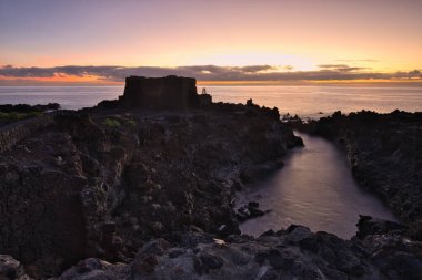Şafak vakti La Palma 'nın engebeli lav kıyısındaki taş bir binanın siluetli kalıntıları. Yumuşak bulutlar ve Atlantik Okyanusu üzerinde pembemsi bir ufuk ile uzun pozlama.