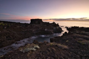 Şafak vakti La Palma 'nın engebeli lav kıyısındaki taş bir binanın siluetli kalıntıları. Yumuşak bulutlar ve Atlantik Okyanusu üzerinde pembemsi bir ufuk ile uzun pozlama.