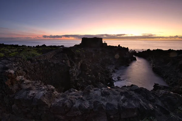 Şafak vakti La Palma 'nın engebeli lav kıyısındaki taş bir binanın siluetli kalıntıları. Yumuşak bulutlar ve Atlantik Okyanusu üzerinde pembemsi bir ufuk ile uzun pozlama.