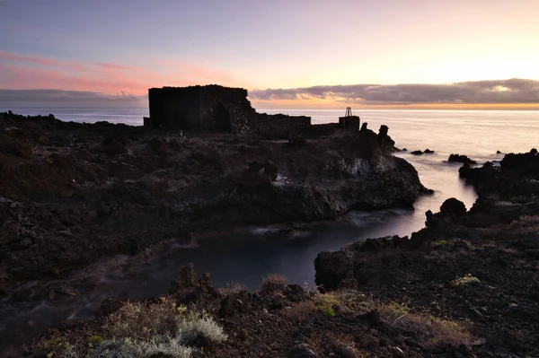 Şafak vakti La Palma 'nın engebeli lav kıyısındaki taş bir binanın siluetli kalıntıları. Yumuşak bulutlar ve Atlantik Okyanusu üzerinde pembemsi bir ufuk ile uzun pozlama.