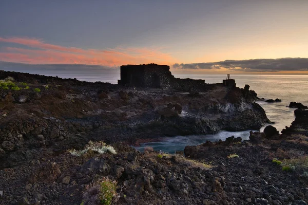 Şafak vakti La Palma 'nın engebeli lav kıyısındaki taş bir binanın siluetli kalıntıları. Yumuşak bulutlar ve Atlantik Okyanusu üzerinde pembemsi bir ufuk ile uzun pozlama.