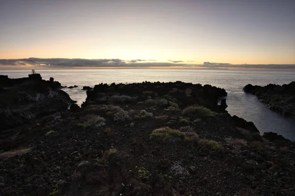 Şafak vakti La Palma 'nın engebeli lav kıyısındaki taş bir binanın siluetli kalıntıları. Yumuşak bulutlar ve Atlantik Okyanusu üzerinde pembemsi bir ufuk ile uzun pozlama.