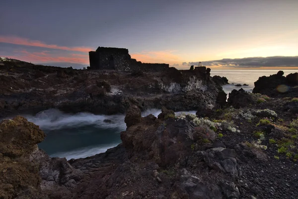 Şafak vakti La Palma 'nın engebeli lav kıyısındaki taş bir binanın siluetli kalıntıları. Yumuşak bulutlar ve Atlantik Okyanusu üzerinde pembemsi bir ufuk ile uzun pozlama.