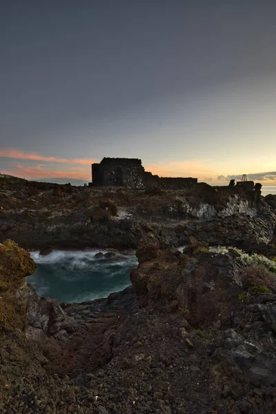 Şafak vakti La Palma 'nın engebeli lav kıyısındaki taş bir binanın siluetli kalıntıları. Yumuşak bulutlar ve Atlantik Okyanusu üzerinde pembemsi bir ufuk ile uzun pozlama.