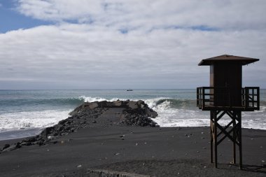 Puerto de Tazacorte, La Palma 'nın kara kumsalındaki ikonik ahşap cankurtaran kulesi. Atlantik dalgaları ve Kanarya Adaları 'ndaki taş iskeletiyle çarpan dramatik volkanik kıyı şeridi..
