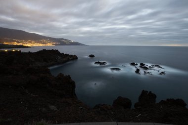 Santa Cruz de La Palma, Kanarya Adaları 'nda uzun süre volkanik kayalık sahile maruz kaldı. Arka planda şehir ışıkları ve Atlantik Okyanusu üzerinde dramatik bulutlu gökyüzü vardı.