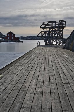 İsveç 'in Skrhamn kentindeki donmuş bir rıhtımda ikonik ahşap dalış platformu. Klasik kırmızı balıkçı kulübesi ve kasvetli kış gökyüzünün altındaki kayalık kıyı şeridi. Kuzey yaşam tarzı ve kış yüzüşü