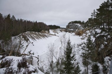 İsveç 'in Fengersfors kentindeki açık taş ocağının panoramik kış manzarası. Zemini kaplayan derin kar ve şafak vakti engin, yumuşak bir kış gökyüzünün altındaki sarp kayalık duvarlar..