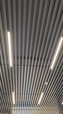 Modern çalışma alanı koridorunda ritmik simetri ve serin atmosfer yaratan doğrusal ışıklarla kaplı dikey eğimli tavan, akıllı