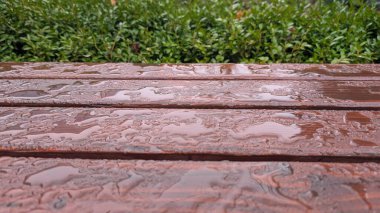 Yağmur damlalı ıslak ahşap bank, su boncukları ve ahşap tahılları gösteren yakın tahta yüzey, bulanık yeşillik arkaplan oluşturan