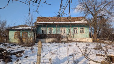 Karla kaplı yeşil çiftlik evi, sarkık çatı çizgileri, soyulmuş boyalar, kırık kepenkler, yapraksız ağaçlar ve yıpranmış çitler.