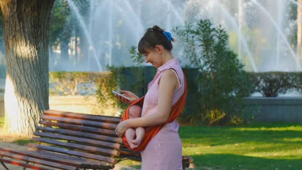Young woman with sleeping baby in sling happily leafs phone gallery — Stock Video