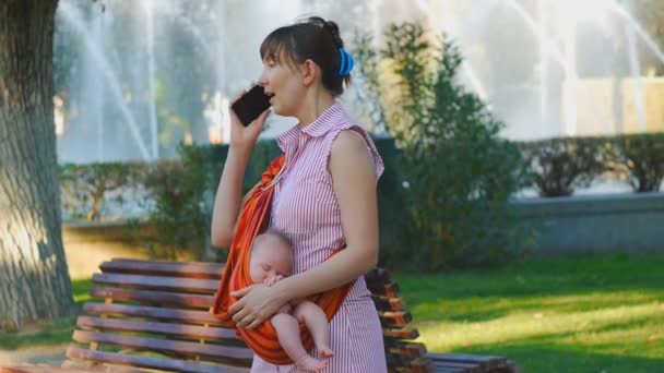 Jonge vrouw met slapende baby in draagdoek begint telefoongesprek — Stockvideo
