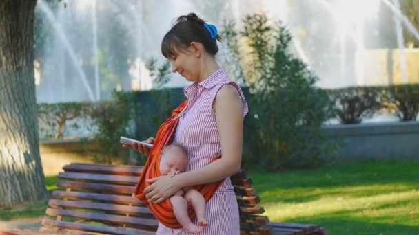 Young woman with sleeping baby in sling smiles using smartphone — Stock Video