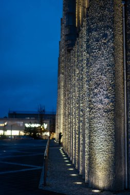 Hallgrimskirkja 'nın desenli beton sütunları aşağıdan aydınlatılır, arka planda sönük şehir ışıklarıyla birlikte masmavi bir gökyüzüne karşı kurulur..