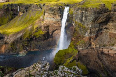 Haifoss waterfall plunges into a deep canyon, surrounded by rugged cliffs, green moss, and rocky terrain, with a faint rainbow near its base.