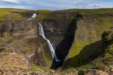 Haifoss waterfall plunges into a deep canyon with basalt cliffs, vibrant green moss, and a smaller waterfall in the background under a clear sky.