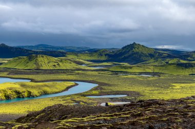Rugged Icelandic landscape with moss covered hills, volcanic rocks, winding blue lakes, rivers, and a prominent peak under an overcast sky.