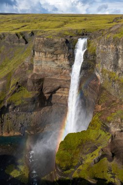 Haifoss waterfall plunges into a deep canyon surrounded by cliffs and green moss, with a rainbow in the mist and a grassy plateau above.