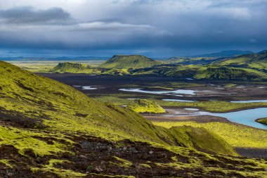 Vibrant green moss covered hills and valleys with volcanic soil, small reflective water bodies, and distant flat topped mountains under a cloudy sky.