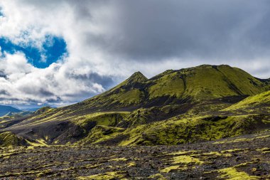Moss covered hills and dark volcanic soil create a vivid contrast under a partly cloudy sky with patches of blue in Iceland's rugged terrain.