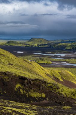 Rolling green moss covered hills contrast with dark volcanic soil, rugged mountains, and scattered water bodies under a cloudy Icelandic sky.