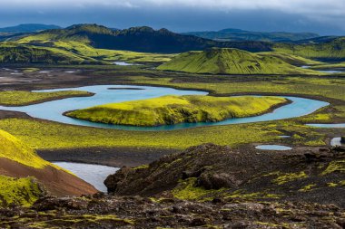 Vibrant green moss covered hills surround bright blue lakes, with volcanic soil and distant mountains under a cloudy sky in Iceland.