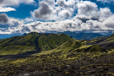 Rolling green moss covered hills and dark volcanic terrain with rugged mountains under a partially cloudy sky in Iceland.