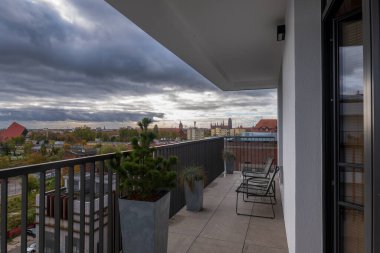 Siyah sandalyeli ve uzun saksılı modern bir balkon Gdansk Eski Kasabası 'na, kırmızı çatılara ve Motlawa Nehri' nin yanındaki kilise kulesine bakıyor..