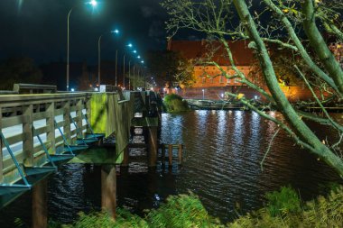Malbork, Polonya 'daki Malbork Kalesi geceleri Nogat Nehri' nin ötesine yükselir. Lambalı bir yaya köprüsü ve sokak lambaları, dalgalı yansımalarla kaleye doğru gidiyor..