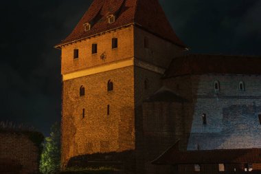 Sıcak spot ışıkları kırmızı tuğla duvarlar, yüksek Gotik bir kule ve Polonya 'nın Malbork Şatosu' nda dik kiremitli çatı. Yandan aydınlatma geceleri derin gölgeler bırakır..