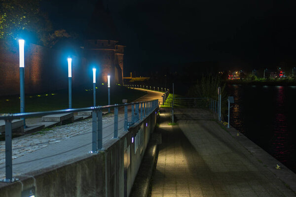 A riverside path with cool blue lights leads to Malbork Castle on the Nogat River in northern Poland. Low key lighting and long perspective emphasize the Gothic walls.