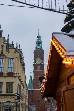 Wroclaw 'daki kış sokağı Rynek' teki Old Town Hall saat kulesini gösteriyor, tuğla cephe ve yeşil kule apartmanlar arasında yükseliyor, kar ve soğuk gün ışığında sıcak kabin ışıkları.