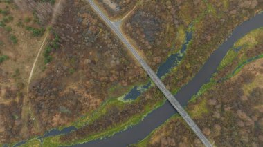 Havadan bakıldığında, Belarus 'ta karanlık dolambaçlı bir nehir boyunca uzanan dar iki şeritli bir köprü, yamalı kış sulamaları, sazlıklar, yosunlu kanallar ve yumuşak ışıkta yapraksız ağaçlar görülüyor..