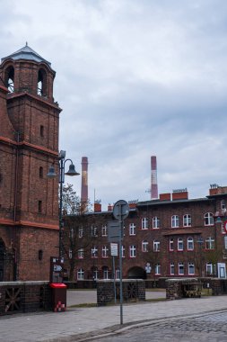 Katowice 'deki Spring Caddesi' nde tuğla işçisi mülkleri, bir kilise kulesi ve ikiz bacalar var. Çakıl taşı kaldırım taşları, işlenmiş tuğla korkuluklar ve maden arabaları bulutlu ışık altında görünür..