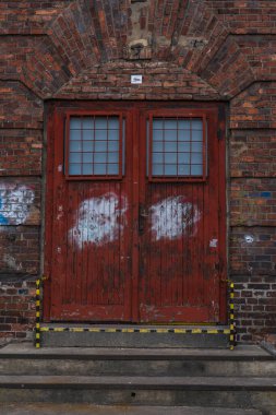 Çitli pencereli yıpranmış kırmızı çift kapı Polonya 'nın Katowice şehrinde tuğla kemerde duruyor. Beyaz boya bantları grafiti kaplıyor, tehlikeli çizgiler işaretler, kapalı bahar ışığı.