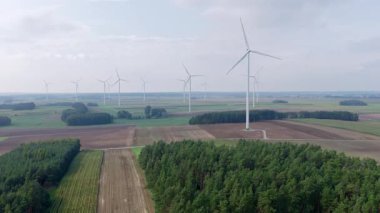 Ekilmiş tarlalar ve kozalaklı tarlalar üzerindeki rüzgar türbinlerinin hava görüntüsü. Bıçaklar hafif rüzgarda döner dağınık gün ışığında ve bulutlu gökyüzünde.
