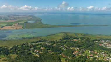 Uzun dar bir tükürük, yamalı tarlalar, sulak alanlarda düz bir kanal ve kıvrımlı bir yol boyunca uzanan sık ağaçlarda kırmızı çatı evleri olan bir kıyı lagününün havadan görünüşü..
