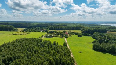 Çayırların ve karışık ormanların ortasındaki tenha bir çiftliğin havadan görünüşü, bahçeli, uzak su, tepeler ve yaz kümülüs bulutlarıyla kırmızı çatı evine uzanan toprak bir yol..