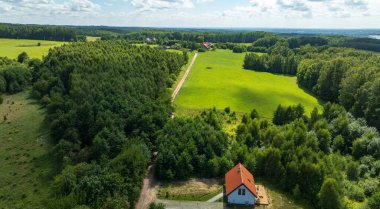 Hava görüntüsü orman, çayır ve tarlaların yanında kırmızı çatılı beyaz bir ev gösteriyor. Bir toprak yol uzak çiftlik evlerine götürür yumuşak gün ışığı ve bulutlar altında.
