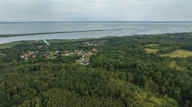 Aerial view shows red and white roofs in dense forest by a calm lagoon. A long breakwater spans the water as a channel cuts inland under soft overcast light.